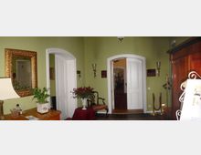 Ein eleganter Eingangsbereich mit hellgrünen Wänden, weißen Doppeltüren und klassischen Holzmöbeln. Ein großer, goldgerahmter Spiegel, Dekorationen wie eine Tischlampe und Blumenarrangements ergänzen das traditionelle Ambiente, während ein Durchgang in einen weiteren Raum mit Teppich führt.