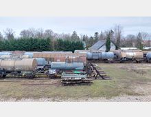 Mehrere Schienentankwagen stehen auf einem Abstellgleis in einer ländlichen Umgebung, umgeben von Bäumen und einzelnen Wohnhäusern. Im Vordergrund sind Schienen und Grasflächen zu sehen, einige Waggons weisen Graffiti auf.