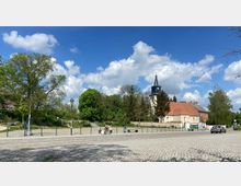 Dorfszene mit einer Kirche mit hellem Mauerwerk und einem spitzen Turm in der Mitte, umgeben von Bäumen und Sträuchern. Im Vordergrund ein gepflasterter Platz mit einem Fußgänger, einer Person im Rollstuhl, sowie parkenden Autos; blauer Himmel mit Wolken im Hintergrund.