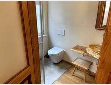 Kleines Badezimmer mit einer freihängenden Toilette, einem Waschtisch aus Holz mit einem steinernen Aufsatzwaschbecken und einem Holzschemel. Der Boden besteht teils aus Holz, teils aus Beton, und im Hintergrund ist ein Fenster sichtbar.