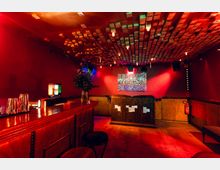 Innenraum einer Bar oder eines kleinen Clubs mit roten Wänden, Holztheke und Barhockern sowie einem DJ-Pult an der Rückwand. Auffällig sind die abgehängte Decke aus vielen Holzquadern, warme Deckenlampen und eine beleuchtete Wandfläche über dem Pult.