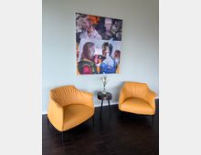 Zwei gelbe Sessel stehen nebeneinander auf dunklem Holzboden, getrennt durch einen kleinen runden Beistelltisch mit einer Vase und einer einzelnen Pflanze. An der mintfarbenen Wand dahinter hängt ein abstraktes Gemälde mit mehreren überlappenden Porträts.