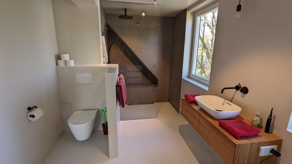 Modernes Badezimmer mit bodengleicher Walk-in-Dusche mit kleinen dunkelgrauen Fliesen und wandhängender Toilette hinter einer halbhohen Trennwand. Rechts ein schwebender Holz-Waschtisch mit ovalem Aufsatzbecken und schwarzer Armatur neben einem großen Fenster.