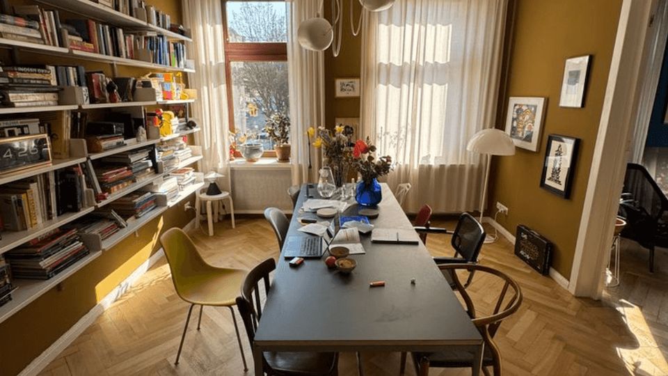 Sunlit dining room with mustard-colored walls, a herringbone wood floor, and a long rectangular table surrounded by mismatched chairs beneath modern pendant lights. A wall-length bookshelf lines one side, and large windows with sheer curtains face the table, with framed artwork and doorways to adjacent rooms on the right.