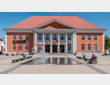 Ein Kulturzentrum mit oranger Fassade und weißen Säulen, zentral gelegen auf einem gepflasterten Platz mit einer flachen Wasserfontäne im Vordergrund. Rechts sind zwei Personen, eine davon im Rollstuhl, zu sehen; links steht ein Restaurant mit Sonnenschirmen.