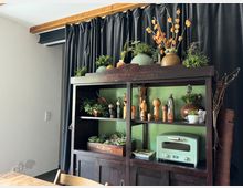 Holzregal im rustikalen Stil vor einem schwarzen Vorhang, dekoriert mit Pflanzen, Tongefäßen, Holzfiguren und anderen kleinen Objekten. Auf der untersten Ebene steht ein hellgrüner Toaster, und darüber sind verschiedene Deko-Elemente auf grünen Regalböden arrangiert.