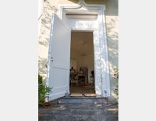 Geöffnete weiße Tür eines Gebäudes mit dekorativem Rahmen, die in einen hellen Raum führt; im Inneren sind eine Kücheninsel, ein Kühlschrank und Holzstühle sichtbar. Der Eingangsbereich ist mit grauen Steinplatten und Matten gestaltet, umgeben von Pflanzen.