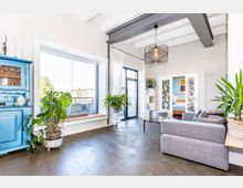 Helles, modernes Wohnzimmer mit großen Fenstern, die viel Tageslicht hereinlassen, grauem Sofa, einer blauen Vitrine, mehreren Zimmerpflanzen und einem Blick auf einen Balkon. Im Hintergrund sind Glastüren mit buntem Muster, die in ein Schlafzimmer führen.