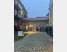 Außenansicht eines gepflasterten Hofes mit umgebenden Wohngebäuden; links Balkone mit Glasgeländern, rechts ein Gebäude mit schlichtem Design und Treppenaufgang. Im Hintergrund ein Torbogen mit erleuchteten Fenstern und überdachtem Durchgang, beleuchtet durch eine Außenlampe.