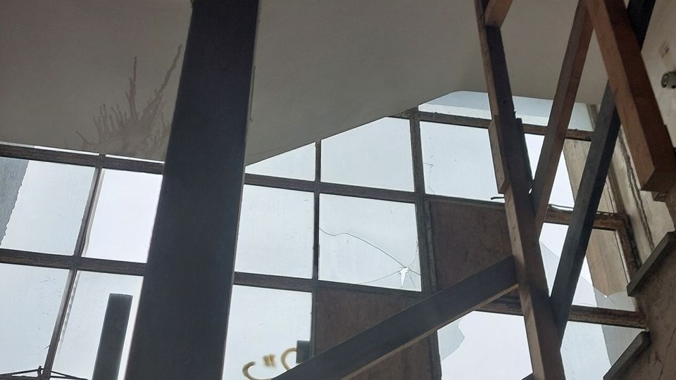 Verlassene Treppe in einem Gebäude mit großen, vergitterten Fenstern und sichtbaren Metall- und Holzelementen; die Umgebung wirkt unfertig oder im Verfall begriffen.