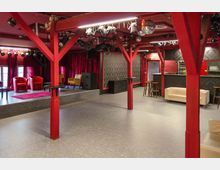 Veranstaltungsraum mit einer kleinen Bühne im Hintergrund, roten Vorhängen und bunten Polstermöbeln. Die Holzbalken im Raum sind rot gestrichen, an der Decke hängen Bühnenscheinwerfer und eine Discokugel, rechts befindet sich eine Bar mit schwarzen Verzierungen.