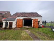 Altes landwirtschaftliches Gebäude mit weiß getünchten Ziegelwänden und einem roten Ziegeldach. Die Scheune hat offene Holztore und wird von einer kleinen, angrenzenden Nebeneinheit mit einer Holztür und einem Fenster ergänzt, umgeben von einer Wiese und leicht bewölktem Himmel im Hintergrund.