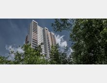 Ein modernes Hochhaus mit weißen und pastellfarbenen Fassadenelementen, umgeben von dichtem, grünem Laub. Der Himmel ist teilweise bewölkt, und Bäume rahmen das Gebäude ein.