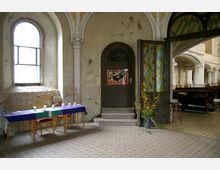 Innenraum einer Kirche mit einer abgenutzten Wand und einem hohen Rundbogenfenster. Ein Tisch mit blauer Decke und weißen Kerzen steht links, daneben eine geschlossene Holztür mit Glasgittern, flankiert von bunten Bleiglasfenstern und einer Vase mit gelben Zweigen. Im Hintergrund sind Kirchenbänke sichtbar.