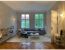 Wohnzimmer in einem Altbau mit Parkettboden, großen Fenstern mit Blick auf grüne Bäume, einem grauen Ecksofa, einem weißen Couchtisch und einer Wandregalablage mit dekorativen Gegenständen. Wände sind in neutralen Tönen gestrichen, und die Beleuchtung besteht aus Wandlampen.