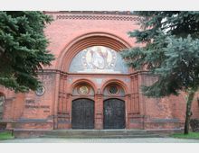 Historische Backsteinkirche mit einem reich verzierten Eingangsportal, bestehend aus zwei dunklen Holztüren, Rundbögen und einem Mosaik mit einer Christusdarstellung. Umgeben von hohen Tannen und einem gepflasterten Vorplatz.