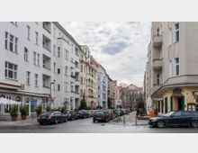 Wohnstraße in einer urbanen Umgebung mit mehrstöckigen Altbauten, die helle Fassaden und Balkone aufweisen. Am Straßenrand parken zahlreiche Autos, und im Hintergrund sind Bäume und ein größeres Gebäude mit roten Dachziegeln zu sehen.