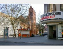 Straßenszene mit einem geschlossenen Imbissstand, einer Werbesäule und einem Bushaltestellenschild in der Nähe eines modernen Gebäudes und einer roten Backsteinkirche im Hintergrund. Ein Geschäft mit einem Werbeschild für einen Sonderverkauf ist rechts im Bild zu sehen.