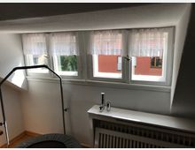 Dachzimmer mit schräger Decke und einer Fensterfront aus drei schmalen Fenstern mit weißen, halbhohen Gardinen. Im Vordergrund ein Trampolin und eine weiße Heizung mit Abdeckung. Im Hintergrund ist ein rotes Gebäude sichtbar.