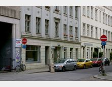 Straßenszene mit mehrstöckigen Gebäuden im Gründerzeitstil, verziert mit Reliefs und Fensterläden; im Vordergrund geparkte Autos, Fahrräder und ein "Durchfahrt verboten"-Schild. Das Erdgeschoss beherbergt Geschäfte und Eingänge, einige Fenster zeigen Blumen.