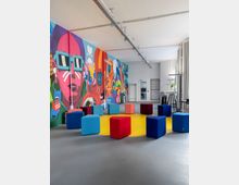 Großer, moderner Veranstaltungs- oder Gemeinschaftsraum mit hoher Decke und sichtbaren Schienen, an einer langen Wand mit farbenfrohem Graffiti-Mural. In der Mitte steht ein Kreis aus bunten Würfelhockern auf einem gelben Rundfeld; am Rand befinden sich Stehtische, Vorhänge und große Fenster.