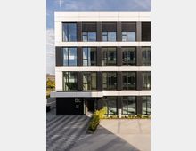 Moderne Bürogebäude-Fassade mit einer Kombination aus weißen und schwarzen Paneelen sowie großen Glasfenstern. Davor eine gepflasterte Fläche mit kleinen bepflanzten Beeten nahe dem Eingang, der mit der Hausnummer "6C" gekennzeichnet ist.