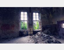 Verlassener Raum mit zwei großen, teils zerbrochenen Fenstern, durch die dichtes grünes Laub zu sehen ist. Die Wände sind beschädigt, teils mit Graffiti versehen, und der Boden ist von Schutt und Trümmern bedeckt.