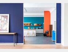 Moderne Küche mit offenen Regalen und weißen Unterschränken, vor einer türkisfarbenen Wand. Im Vordergrund ist ein Durchgang mit einer dunkelblauen Wand und einer Holzkonsole zu sehen, rechts eine orangefarbene Wand als Akzent.