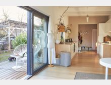 Helle, moderne Wohnküche mit Holzfußboden, offener Küchenzeile und großen Schiebetüren, die auf eine Terrasse mit weißen Stühlen und Pflanzen führen. Im Raum stehen dekorative Elemente wie eine Schneiderpuppe und Körbe mit Zickzack-Muster.