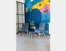 Sitzecke in einem modernen Innenraum mit zwei grauen Lounge-Sesseln, passenden Fußhockern und kleinen Beistelltischen auf glattem Boden. Im Hintergrund dominiert eine großflächige, farbenfrohe Wandmalerei mit abstraktem Gesichtsmotiv sowie ein Fenster mit Vorhang und ein Heizkörper.