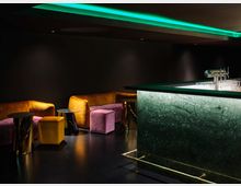 Eine stilvolle Bar mit einem Tresen aus grünem Marmor, beleuchtet durch dezente Lichter. Im Hintergrund befinden sich Sitzbereiche mit gelben und violetten Polstern sowie kleinen, metallischen Beistelltischen.