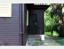 Dunkelbraunes Holzhäuschen mit einer schmalen schwarzen Tür, die über ein paar Betontreppenstufen mit Metallgeländer erreichbar ist. Im Hintergrund befinden sich dichte grüne Bäume und Sträucher.