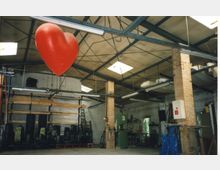 Industrielle Werkhalle mit Backsteinpfeilern, Stahlträgern und Oberlichtern. Im Vordergrund hängt ein großes, rotes Herz von der Decke, während im Hintergrund Regale mit Holzmaterialien und eine Wand mit einer Silhouette von Hochhäusern zu sehen sind.