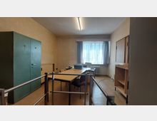 Ein einfacher Büroraum mit einem großen grünen Metallschrank, einem rechteckigen Holztisch mit mehreren Stühlen und einer Schreibmaschine darauf. Im Hintergrund befindet sich ein Fenster mit Vorhängen, ein Heizkörper und weitere Regale und Schränke aus Holz.