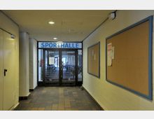 Flur mit hellen Backsteinwänden und Kork-Pinnwänden an den Seiten, der zu einer Glastür mit der Beschriftung „Sporthalle“ führt; der Boden besteht aus dunklen Fliesen.
