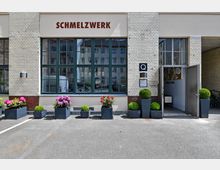 Fassade eines Gebäudes mit hellen Backsteinen und grünen Fenstern, beschriftet mit „SCHMELZWERK”. Vor dem Eingang stehen zahlreiche Blumenkübel mit grünen Buchsbaumkugeln und blühenden Hortensien.