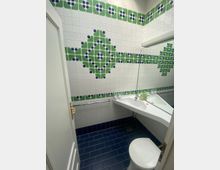 Kleines Badezimmer mit weiß gefliesten Wänden, dekoriert mit grünen und blauen Mosaikmustern. Der Boden ist mit dunkelblauen Fliesen versehen, und es gibt ein weißes Waschbecken mit einem Spiegel sowie eine Toilette im Vordergrund.