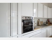 Moderne Küche mit weißen Hochglanzfronten, einem integrierten Backofen, Edelstahlgriffen und einer Arbeitsplatte aus hellem Holz. Daneben befinden sich eine Edelstahlspüle, Küchenutensilien, Schneidebretter und verschiedene Geräte.