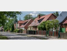 Eine ruhige Wohnstraße mit freistehenden Einfamilienhäusern aus rotem Ziegelstein und Ziegeldächern, flankiert von Zäunen und Bäumen. Im Hintergrund ist die Spitze eines Kirchturms zwischen den Bäumen sichtbar.