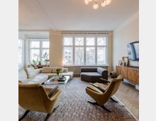 Helles Wohnzimmer mit großen Fenstern, die viel Tageslicht hereinlassen, einer Sitzecke mit Sofa und Sesseln, einem Teppich auf Holzboden und einer Kommode mit einem an der Wand montierten Fernseher. Pflanzen und dekorative Elemente sind dezent verteilt.