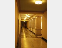 Ein beleuchteter Innenflur mit hellen Wänden, einer Decke mit runden Lampen und einem Boden aus polierten Marmorfliesen. Der Flur ist durch Säulen aus beigefarbenem Stein teilweise unterteilt und führt in die Tiefe.