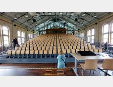 Großer Vortragssaal mit ansteigenden Sitzreihen aus hellem Holz, umgeben von gelben Ziegelwänden mit hohen Rundbogenfenstern. Der Raum hat eine offene Dachkonstruktion mit sichtbaren Stahlträgern und Deckenleuchten.