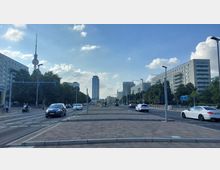Breite Straße in einer städtischen Umgebung mit Autos und Bäumen auf beiden Seiten, flankiert von modernistischen Wohngebäuden. Im Hintergrund ist der Berliner Fernsehturm sowie ein Hochhaus zu sehen, und eine U-Bahn-Station liegt rechts im Bild.