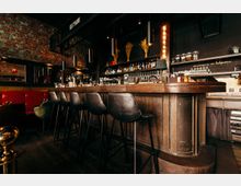 Innenansicht einer Bar im Industrial-Stil mit langer dunkler Holztheke, mehreren hohen Barhockern und Regalen voller Flaschen und Gläser. Sichtmauerwerk aus Ziegeln, schwarze Wände und hängende Zylinderleuchten prägen den Raum.