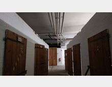 Ein Kellerflur mit weißen Backsteinwänden und mehreren hölzernen Türen auf beiden Seiten. An der Decke verlaufen offene Rohre, und im Hintergrund befindet sich eine Doppeltür aus Holz sowie ein Feuerlöscher an der Wand.