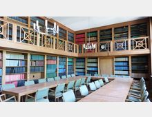 Ein großzügiger Bibliotheksraum mit hölzernen Bücherregalen an allen Wänden, gefüllt mit bunt gebundenen Büchern. In der Mitte stehen lange Holztische mit gepolsterten Stühlen, und zwei moderne Kronleuchter hängen von der Decke.