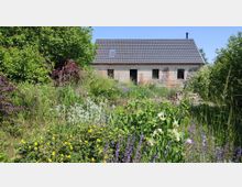 Ein kleines Backsteinhaus mit dunklem Ziegeldach steht inmitten eines üppigen, blühenden Gartens mit hohem Gras, bunten Blumen und Sträuchern, umgeben von grüner Vegetation. Im Hintergrund sind teilweise abgebröckelte Stellen an der Fassade erkennbar.