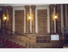 Innenraum mit einer traditionellen Architektur, bestehend aus mehreren Säulen aus braunem Stein und Wandverkleidungen mit rechteckigen Verzierungen. Links führt eine Treppe mit rotem Teppich und einem Metallgeländer nach oben, während Wandlampen warmes Licht spenden.