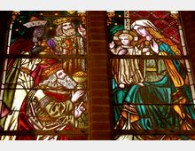 Buntglasfenster in einer Kirche, das eine biblische Szene zeigt: Drei Könige überreichen Geschenke, während Maria mit dem Jesuskind sitzt. Die Szene ist mit detaillierten Mustern und leuchtenden Farben gestaltet.
