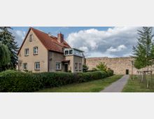 Wohnhaus mit Satteldach und beigefarbener Fassade, umgeben von einer Hecke und Rasenfläche. Im Hintergrund ist eine historische Steinmauer sowie ein gepflasterter Gehweg mit Bäumen sichtbar.
